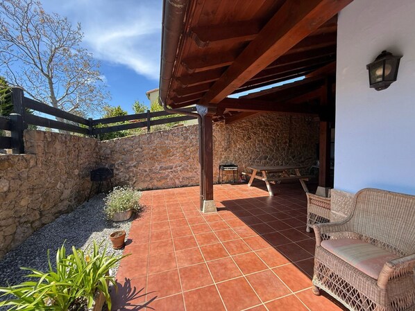 A covered patio with a stone wall, a wooden table, and a wicker chair. - Casa La Escuelina en Villanueva (Villanueva)