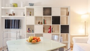 A living room with a glass-top table, a white sofa, a white bookshelf, and a flat-screen TV mounted on the wall. - GuestHost - Arezzo Equestrian Centre-Apt W/ Private Parking! (Arezzo)