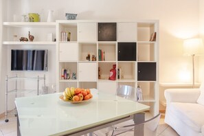 A living room with a glass-top table, a white sofa, a white bookshelf, and a flat-screen TV mounted on the wall.