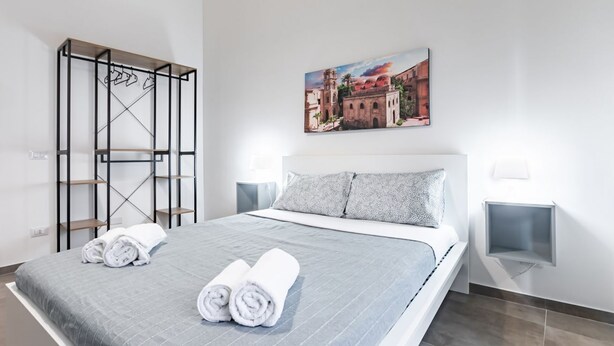 A neatly made bed with rolled towels, a painting of a building, and a metal rack with hangers. - GuestHost - Palazzo dei Normanni & Ballarò Roomy Modern Apt x9 (Palermo)