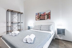 A neatly made bed with rolled towels, a painting of a building, and a metal rack with hangers.