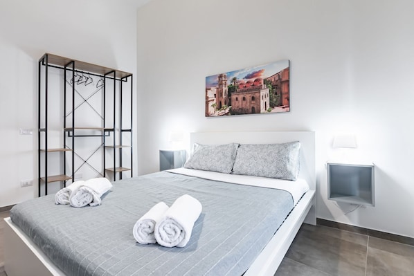 A neatly made bed with rolled towels, a painting of a building, and a metal rack with hangers.