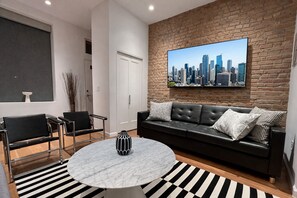 Un salon moderne avec un canapé en cuir noir, une table basse ronde en marbre et un tableau représentant un paysage urbain accroché au mur en briques.