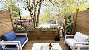 Eine Terrasse im Freien mit Holzboden, weißen und blauen Lounge-Stühlen, einem weißen Tisch mit einer Flasche und Gläsern sowie Pflanzgefäßen mit Grünpflanzen.