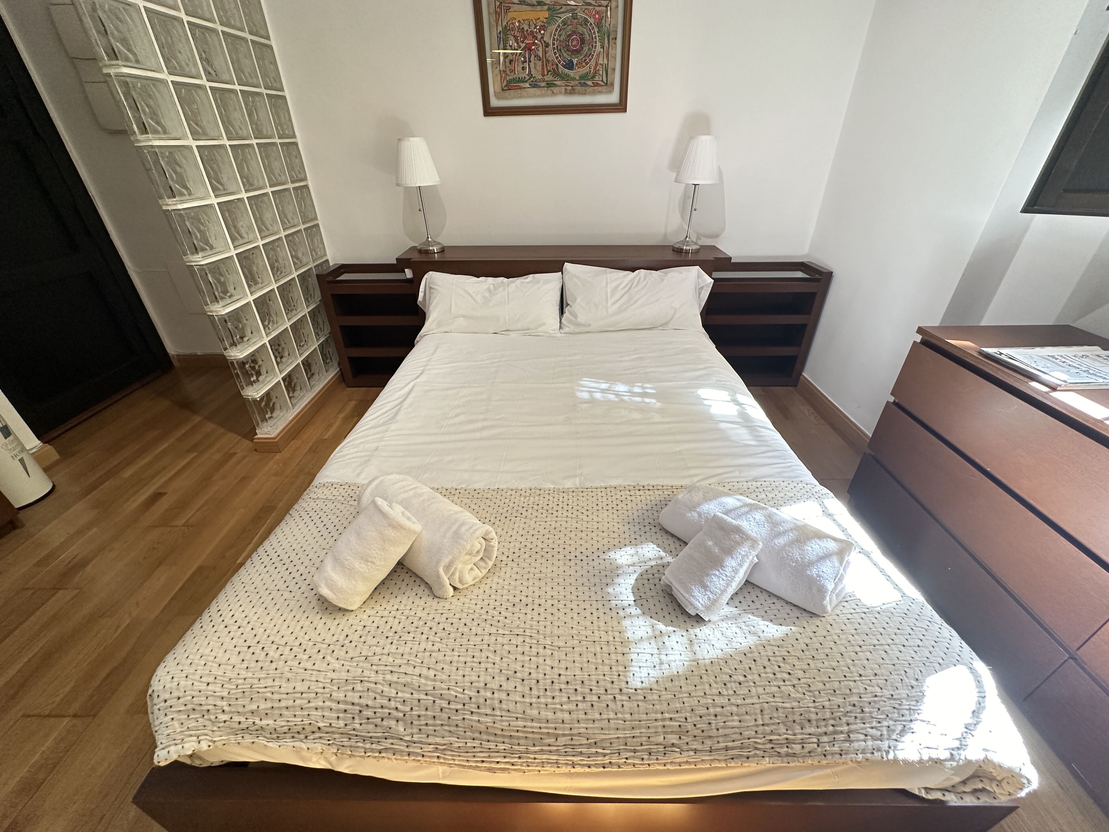 A neatly made bed with white linens and a textured blanket, two bedside tables with lamps, a wooden dresser, and a wall with a decorative piece.