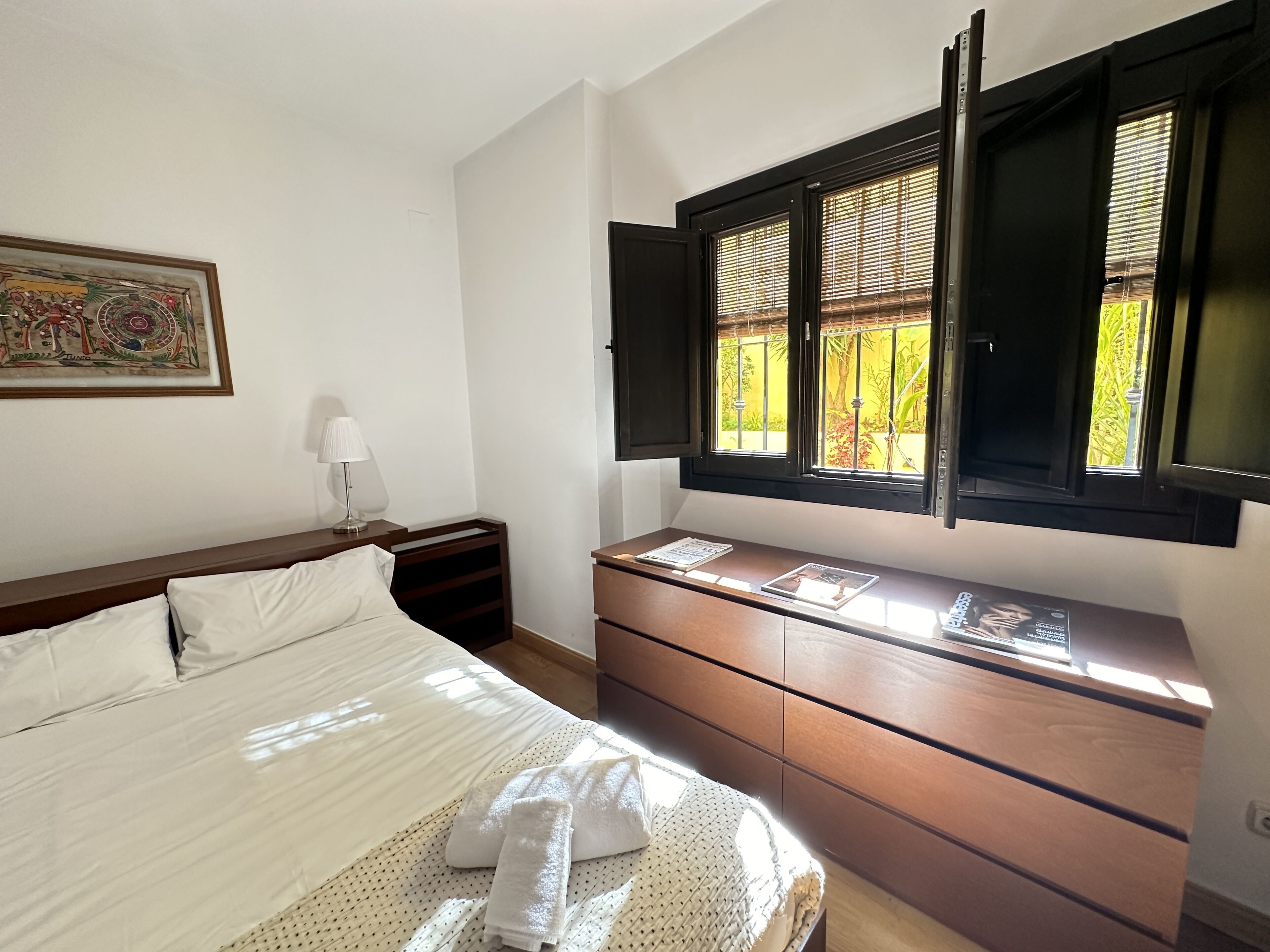 Een slaapkamer met een bed, nachtkastje, dressoir en een raam met geopende luiken.