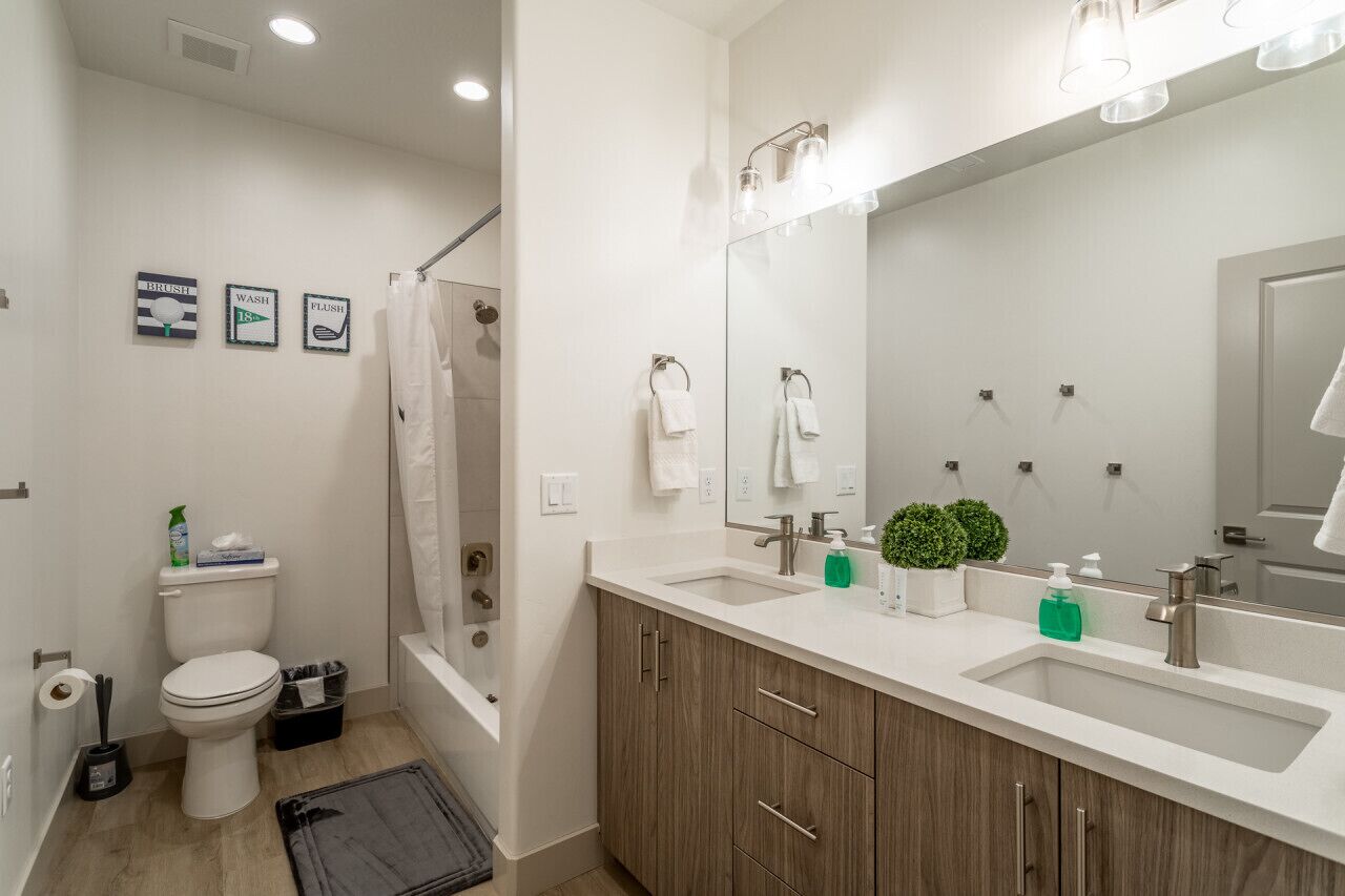 Ein modernes Badezimmer mit Toilette, Dusche und Waschbecken. An der Wand hängen drei gerahmte Schilder, ein großes Spiegel und ein Blumentopf auf der Ablagefläche des Waschbeckens.