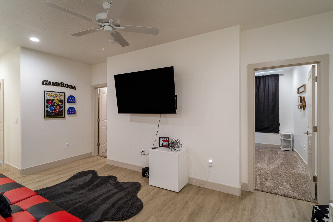 Una sala de estar con ventilador de techo, televisor montado, letrero que indica sala de juegos y un sofá rojo y negro.