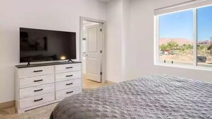 Un dormitorio con cama, televisor sobre una cómoda, ventana con vista a un paisaje desértico y puerta que da a otra habitación.