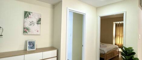 A hallway leading to a bedroom with a bed, a bedside table, and a potted plant.