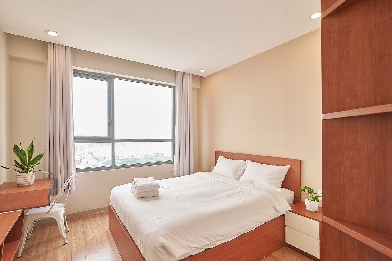A modern hotel room with a large window, a wooden bed, a desk with a chair, and a small potted plant.