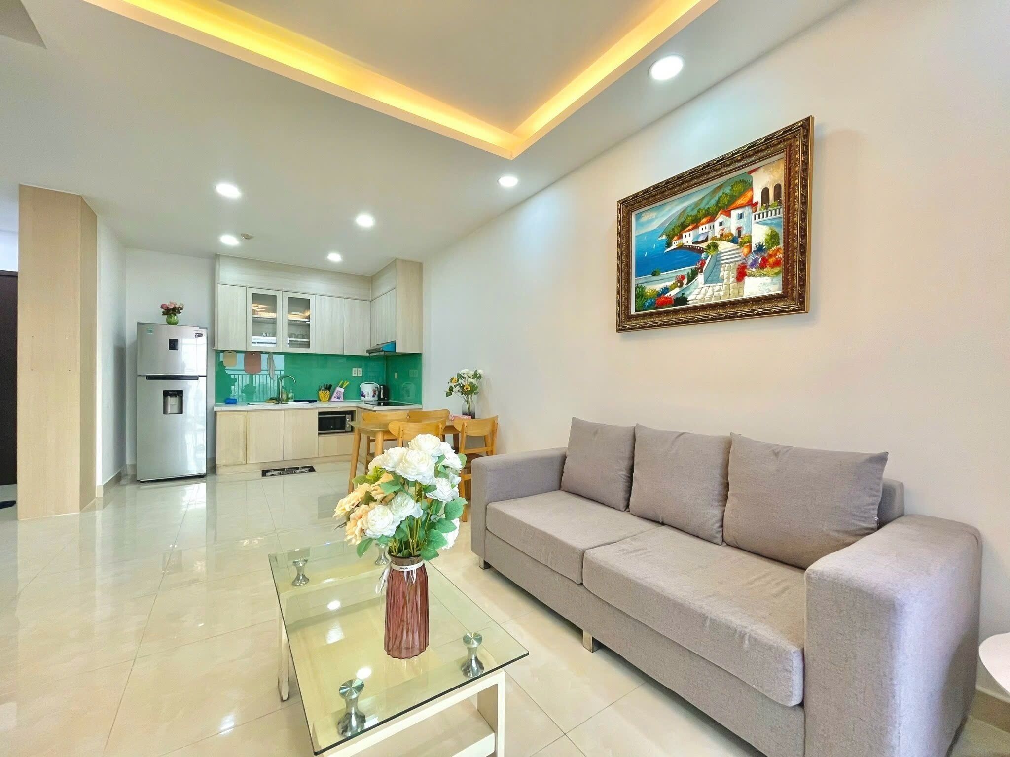 A modern living room with a grey sofa, a glass coffee table with a vase of flowers, a kitchen in the background, and a framed painting on the wall.