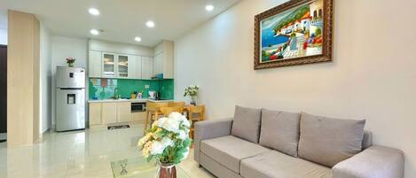 A modern living room with a grey sofa, a glass coffee table with a vase of flowers, a kitchen in the background, and a framed painting on the wall.