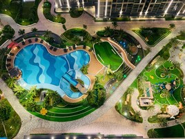 Uma piscina grande com efeito de ondas, cercada por jardins bem cuidados e palmeiras.
