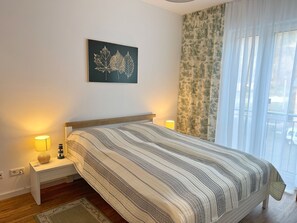 Un dormitorio con cama, lámparas de noche, mesita de noche, alfombra y ventana con cortinas.