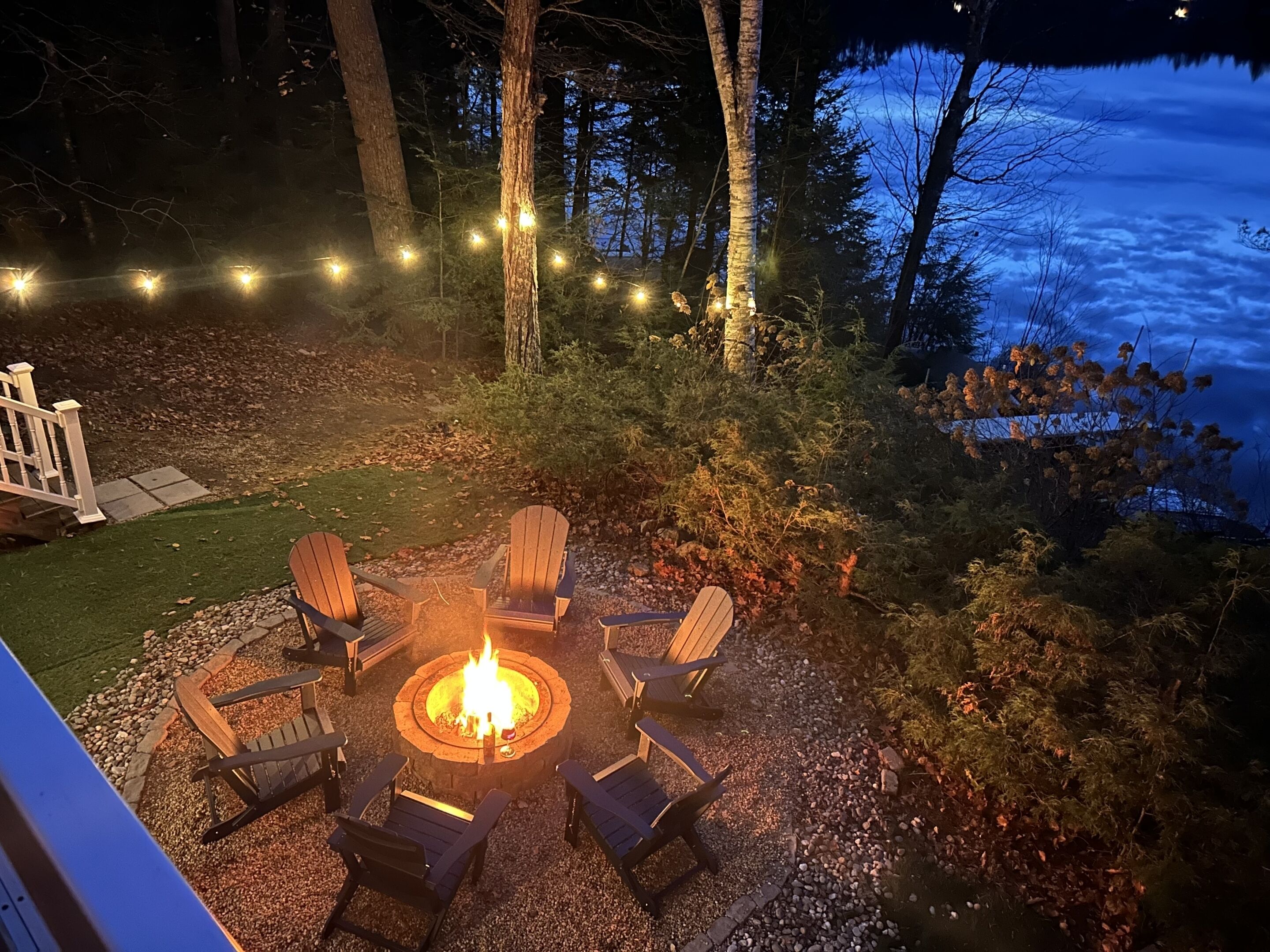 En eldstad omgiven av Adirondack-stolar på en uteplats i en trädgård, med en terrass och ljusslingor.