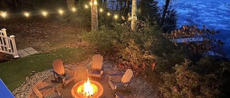 A fire pit surrounded by Adirondack chairs in a backyard with a deck and string lights.