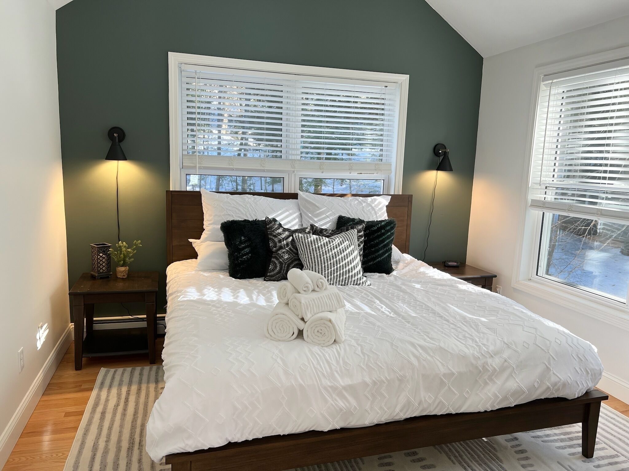 A bedroom with a bed, two bedside tables, a nightstand, a window with blinds, and a lamp on each side.