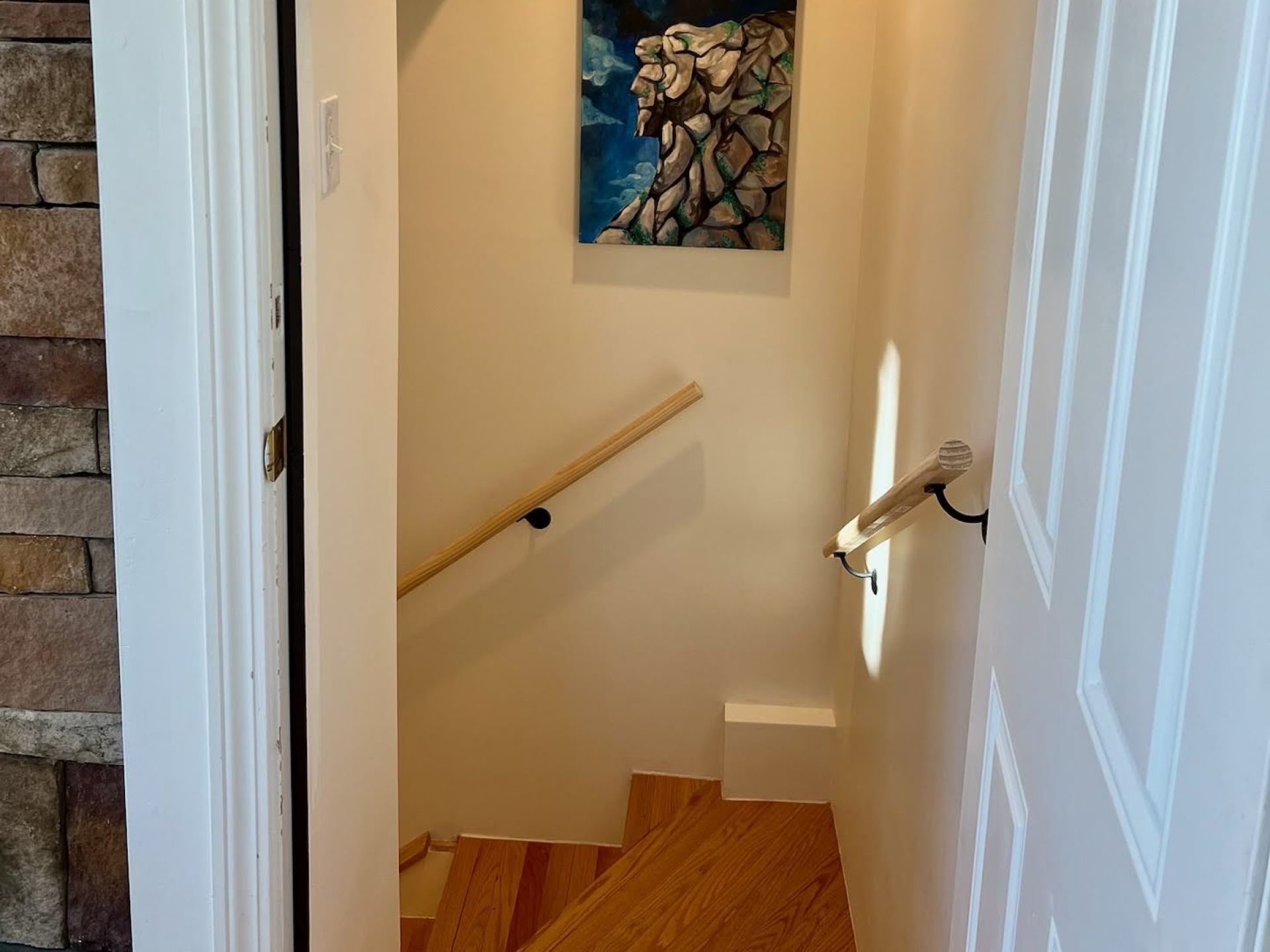 Een trap met houten treden en een leuning, die leidt naar een kamer met een stenen muur en een schilderij van een berg.