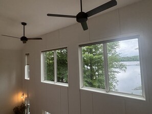 Une pièce dotée de deux ventilateurs de plafond, de grandes fenêtres offrant une vue sur les arbres et un lac, et d’un luminaire fixé au mur.