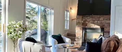 A spacious living room with a fireplace, large windows, and a flat-screen TV mounted on the wall.