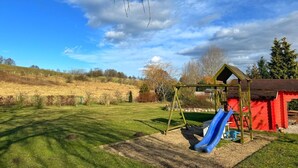 Une aire de jeux avec un toboggan et des balançoires, entourée d’un champ d’herbe et d’un bâtiment rouge.