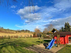 A playground with a slide and swings, surrounded by a grassy field and a red building.
