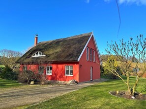 Een rode woning met rieten dak, omgeven door een verzorgd gazon en een kleine boom.