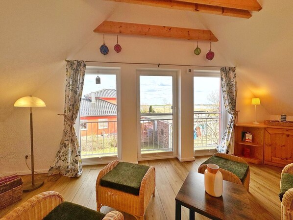 A cozy room with a wooden ceiling, a dining table with chairs, a floor lamp, and a view of a house through large windows.