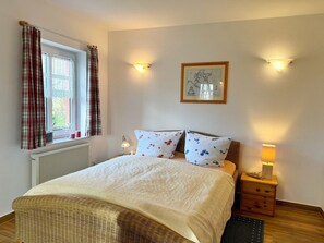A bedroom with a bed, two pillows, a bedside table with a lamp, a framed picture on the wall, and a window with curtains.