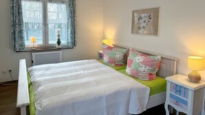 Une chambre à coucher avec un lit, deux tables de chevet avec des lampes, une fenêtre avec des rideaux, un radiateur et un tableau accroché au mur.