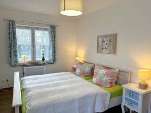 A bedroom with a bed, two bedside tables with lamps, a window with curtains, a radiator, and a wall-mounted picture.