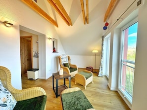 Een knus loft-appartement met hoge plafonds, houten balken, een groot raam, rieten meubels en een kleine tafel.