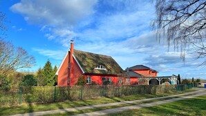 Une maison rouge avec un toit de chaume et une cheminée blanche, entourée d’une clôture verte et d’arbres.