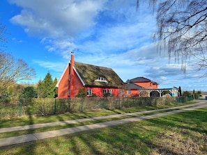 Een rode woning met rieten dak en een witte schoorsteen, omgeven door een groene hekken en bomen.