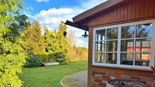 Une maison en bois avec une grande fenêtre, un petit jardin avec une pelouse et un tonneau en bois.