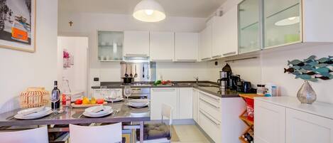 Une cuisine moderne avec des meubles blancs, un coin repas avec des chaises, un luminaire suspendu et une décoration murale représentant des poissons.