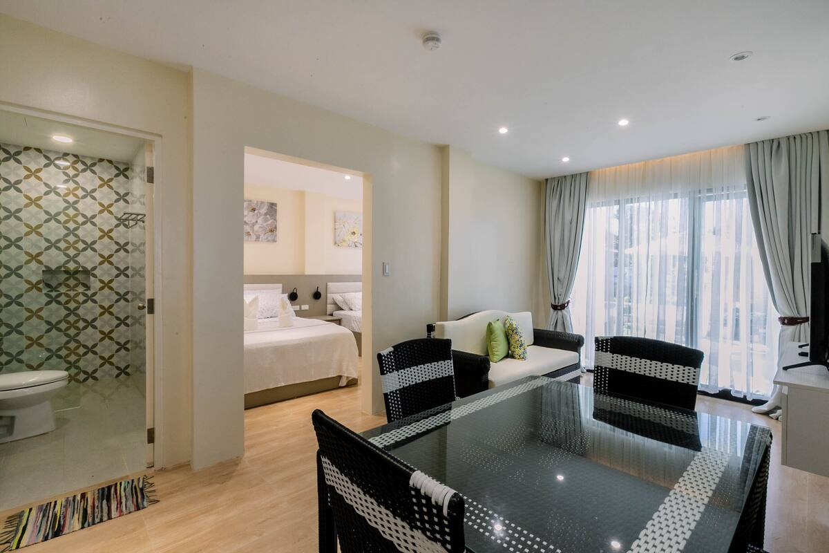 A modern hotel room with a glass shower enclosure, a separate bedroom, a dining area with a glass table, and a living area with a sofa and curtains.