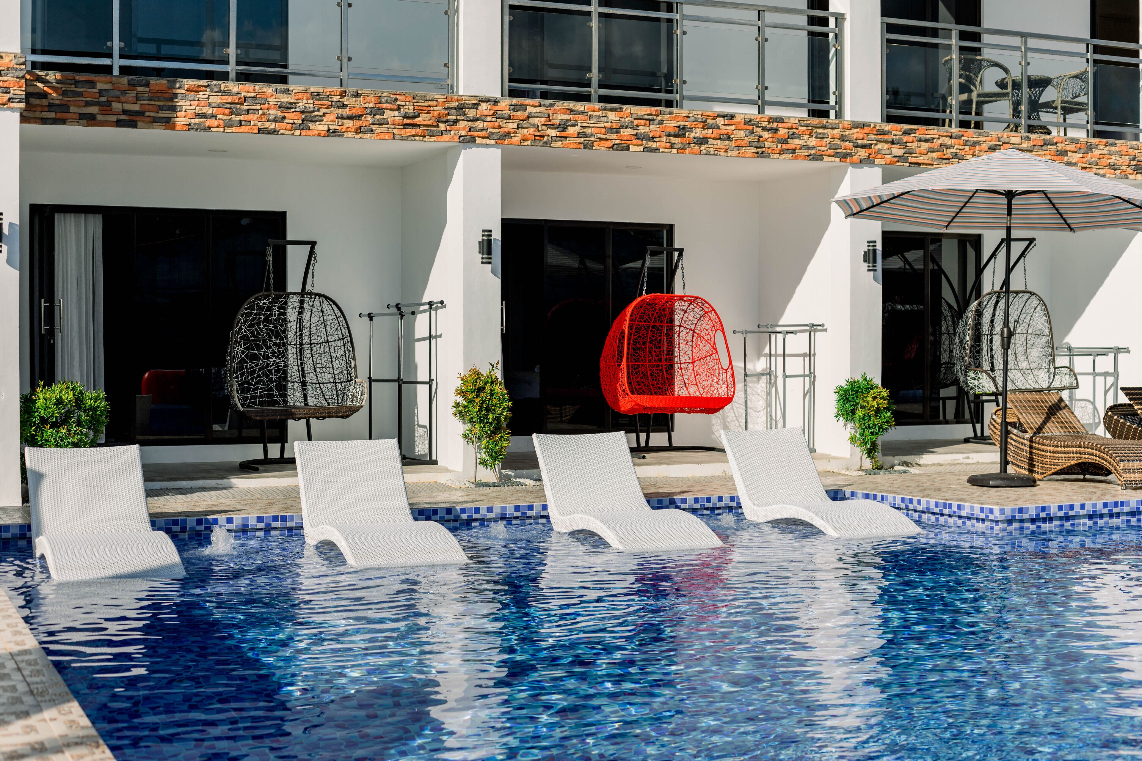 A swimming pool with lounge chairs and a red chair, surrounded by a building with a tiled roof and white walls.