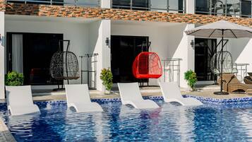 A swimming pool with lounge chairs and a red chair, surrounded by a building with a tiled roof and white walls.