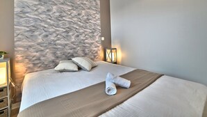 A hotel room with a large textured wall, a bed with white linens, two pillows, a rolled towel, a bedside table with a lamp, and a wall-mounted light fixture. - La Parie 1 by Interhome (Ile d'Oléron)
