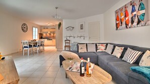 A modern living room with a grey sofa, wooden coffee table, and a dining area with a wooden table and chairs. There is a kitchen in the background with wooden cabinets and a hanging light fixture. - La Parie 1 by Interhome (Ile d'Oléron)