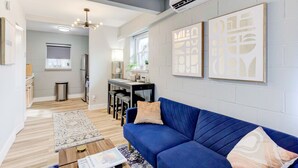 A modern living room with a blue sofa, a coffee table, and a rug. There is a dining area with a table and chairs, and a kitchen area with cabinets and a refrigerator. - Modern Luxe Loft with King Bed, Free Parking & Wi-Fi (Memphis)