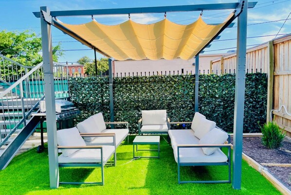 Un área de estar al aire libre con techo, con sillas y una mesa blancas, rodeada por un seto y una valla de madera.