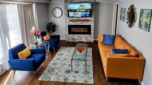 A modern living room with a fireplace, blue and orange sofas, a TV mounted on the wall, and decorative wall art.