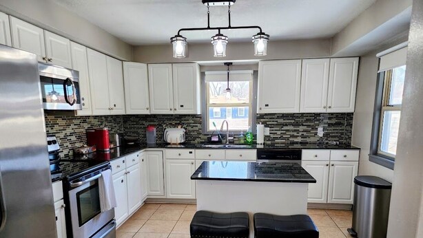 A modern kitchen with white cabinets, a black countertop, a stainless steel refrigerator, a double oven, and a center island with stools.