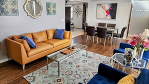 A modern living room with a leather sofa, a glass coffee table, and a blue armchair. There is a dining area with a table and chairs, and a kitchen area with a refrigerator.