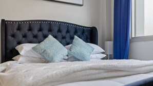 Una cama con cabecera capitonada, ropa de cama blanca y dos almohadas estampadas.