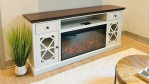 Una sala de estar con una televisión de pantalla plana montada en la pared, un mueble de madera para entretenimiento con chimenea integrada, una mesa de centro redonda y una planta en maceta.