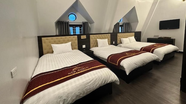 Habitación de hotel con tres camas, cada una con cabecera y colcha. Hay un televisor de pantalla plana montado en la pared y una ventana con vistas al exterior.